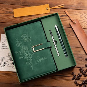 May include: A dark green journal with a floral line art design and the name Sophia engraved on the cover, featuring a gold metal clasp. The set includes a matching green box, a black and silver pen, a pen refill, and two leather bookmarks.