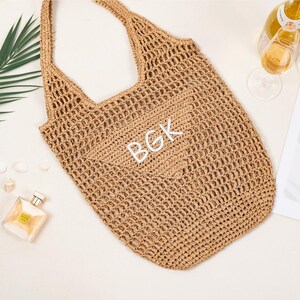 May include: A tan crocheted tote bag with the initials "BGK" in white. The bag has a shoulder strap and a large open weave design. The image also includes a bottle of wine, two glasses, heart-shaped sunglasses, and a perfume bottle.