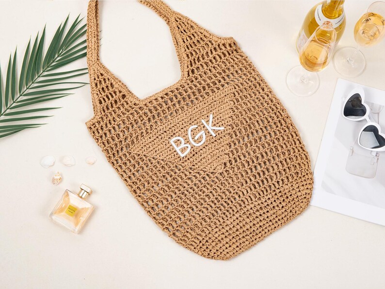 May include: A tan crocheted tote bag with the initials "BGK" in white. The bag has a shoulder strap and a large open weave design. The image also includes a bottle of wine, two glasses, heart-shaped sunglasses, and a perfume bottle.