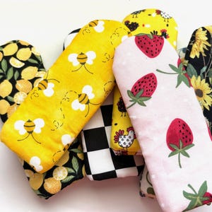 May include: A collection of oven mitts with various patterns. Designs include lemons, bees, strawberries, sunflowers, chickens, and a black and white checkerboard. The mitts are made of fabric and are in various shades of yellow, pink, black, and white.