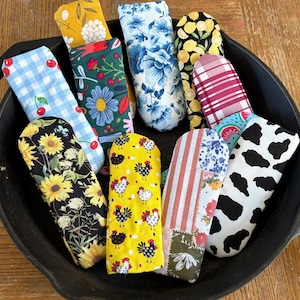 May include: A collection of colorful pot holders in a cast iron skillet. The pot holders feature various patterns, including sunflowers, chickens, floral designs, and a cow print. The pot holders are made of fabric and are designed to protect hands from heat.