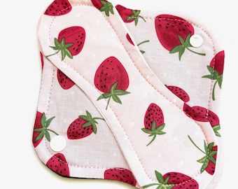 May include: Reusable menstrual pads with a strawberry pattern. The pads feature a light pink background with white polka dots and red strawberries with green leaves. Each pad has a white snap closure.