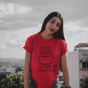 May include: Red t-shirt with rolled sleeves featuring a black graphic of a fishbowl and the text "I'M LIKE A FISH OUT OF WATER WITHOUT GOD". The shirt is paired with blue jeans.