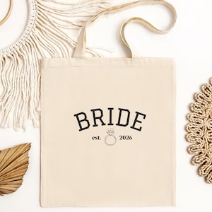 May include: A beige canvas tote bag with the word "BRIDE" in bold black letters. Below the word "BRIDE" is the text "est. 2026" with a diamond ring graphic. The bag has a simple design and is suitable for carrying various items.