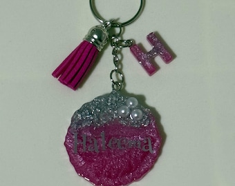 May include: A pink and silver keychain with a tassel, letter "H", and a circular charm. The charm has the name "Haleema" printed on it. The keychain is attached to a silver key ring.