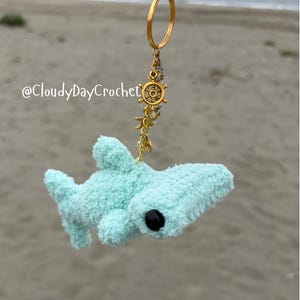 May include: A light blue, plush shark keychain with a gold-colored keyring and nautical charms. The shark has a black eye and a textured, fuzzy appearance. The keychain includes a ship's wheel charm and other sea-themed embellishments.
