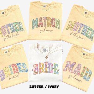 May include: Cream-colored t-shirts with floral text designs for a wedding party. The shirts say "Mother of the Bride," "Matron of Honor," "Mother of the Groom," "Bridesmaid," "Bride," and "Maid of Honor."