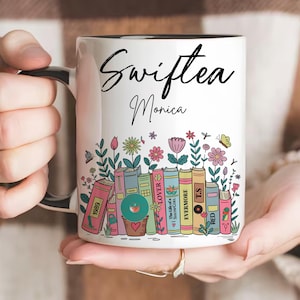 May include: White ceramic mug with a black handle and rim, featuring the text "Swiftiea Monica" in black script. The mug is decorated with colorful flowers, butterflies, and a row of books with titles like "1989" and "Lover".
