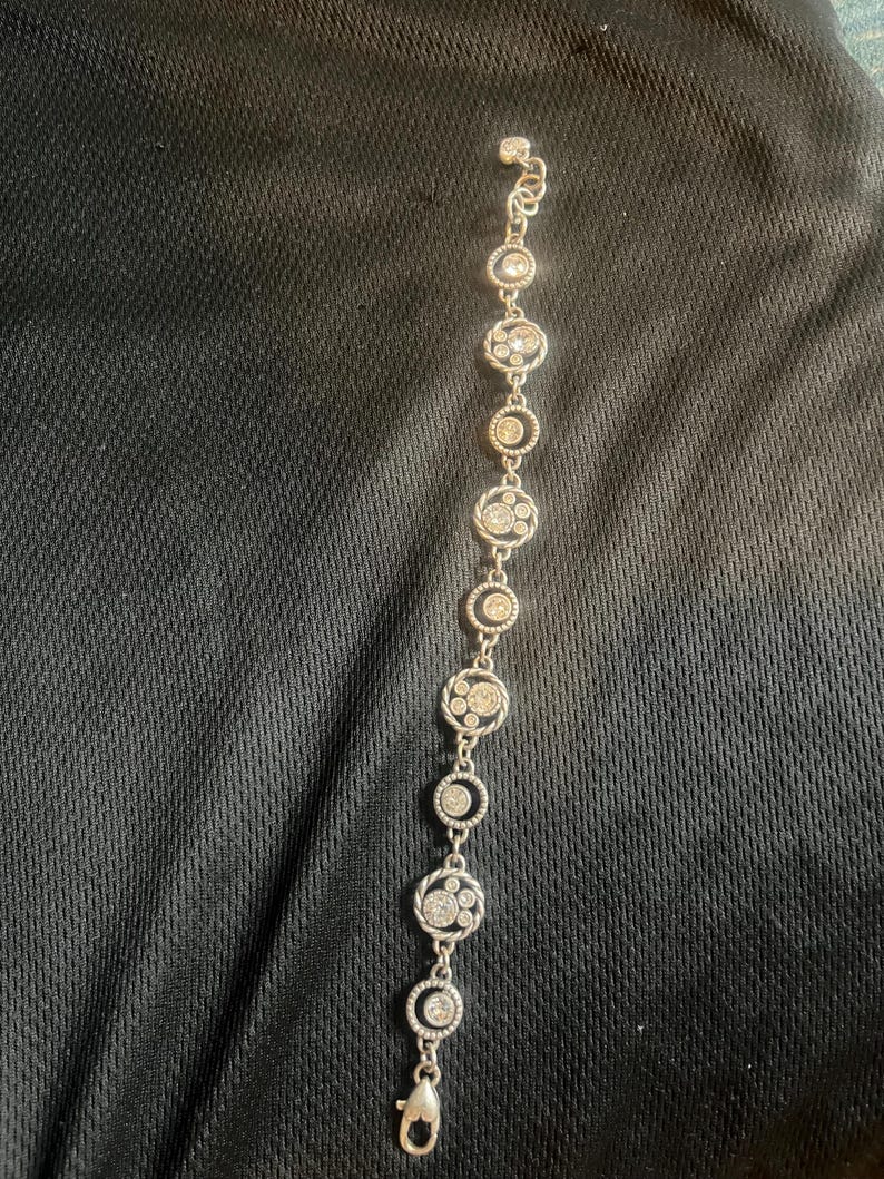 May include: Silver bracelet with a series of circular links. Each link is decorated with clear, sparkling crystals, creating a delicate design. The bracelet has a lobster clasp closure.