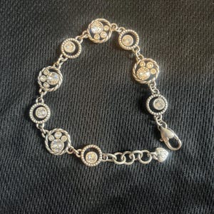 May include: Silver bracelet with circular and paw-shaped links, each set with clear crystals. The bracelet has a lobster clasp and is shown against a dark background. The bracelet's design is elegant.