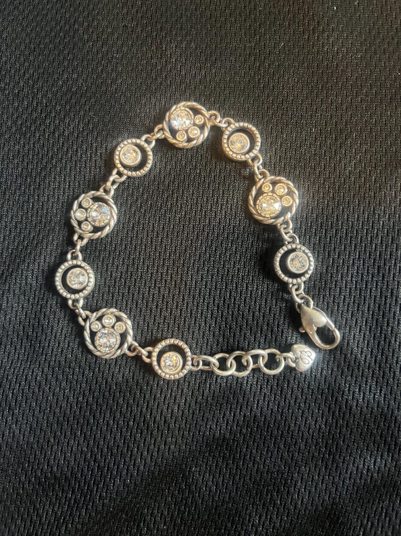 May include: Silver bracelet with circular and paw-shaped links, each set with clear crystals. The bracelet has a lobster clasp and is shown against a dark background. The bracelet's design is elegant.