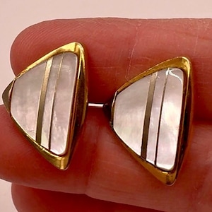 May include: Gold-toned cufflinks with a triangular design. Each cufflink has a white, pearlescent centre with three vertical gold stripes and a gold border. The cufflinks are held by a person's fingers.