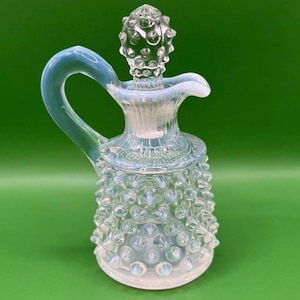 Puede incluir: Una aceitera de vidrio transparente con asa y tapón. La aceitera tiene un diseño texturizado de clavos y un tinte azul claro. El tapón también es transparente y tiene una parte superior redondeada y texturizada. La aceitera está sobre un fondo verde.