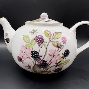 Puede incluir: Tetera de cerámica blanca con tapa, con un diseño floral de flores rosas, hojas verdes y moras. La tetera tiene un asa y un pico curvos, con detalles dorados. El diseño se repite en la tapa.