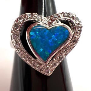 Könnte beinhalten: Ein silberner Ring mit einem doppelten Herzdesign. Das innere Herz ist mit einem leuchtend blauen Opal gefüllt, während das äußere Herz mit kleinen, klaren Edelsteinen besetzt ist. Der Ring wird auf einem schwarzen Ständer präsentiert.