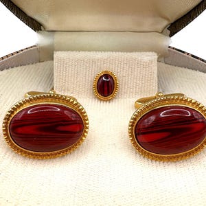 Peut inclure: Un ensemble vintage de boutons de manchette et de pince à cravate dorés. Les boutons de manchette et la pince à cravate présentent des pierres ovales marbrées rouges et noires serties dans un cadre doré décoratif. L'ensemble est présenté dans un coffret marron et crème.