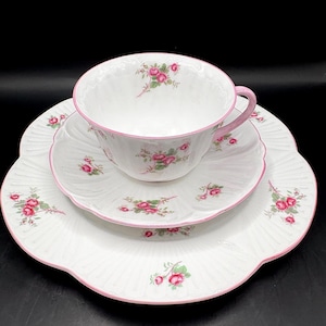 Op de afbeelding: Witte theeservies met roze rozenpatroon en roze rand. De set bestaat uit een kopje, een schotel en een bord. Het kopje heeft een roze oor en het bord heeft een geschulpte rand.