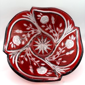 May include: A deep red glass bowl featuring clear cut-glass floral patterns of roses and leaves. The center of the bowl displays a prominent starburst design, while the outer rim has a scalloped edge. The glass transitions from clear to red.