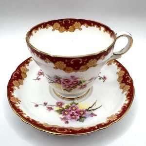 May include: A vintage teacup and saucer set. The white porcelain cup and saucer feature a maroon and gold floral design around the rims. The cup and saucer have a floral pattern with pink flowers and green leaves.