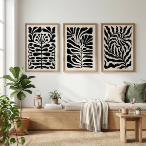 May include: Three framed art prints with abstract black and white botanical designs. The prints are in light wood frames and hang on a white wall. The room is decorated with plants, a wooden bench, and a small side table.