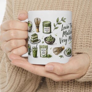 May include: White ceramic mug featuring matcha-themed illustrations, including a whisk, powder, and cake. The mug has the text "Lovin' The Matcha Very Matcha" in a handwritten style. The mug is held by a person wearing a beige sweater.