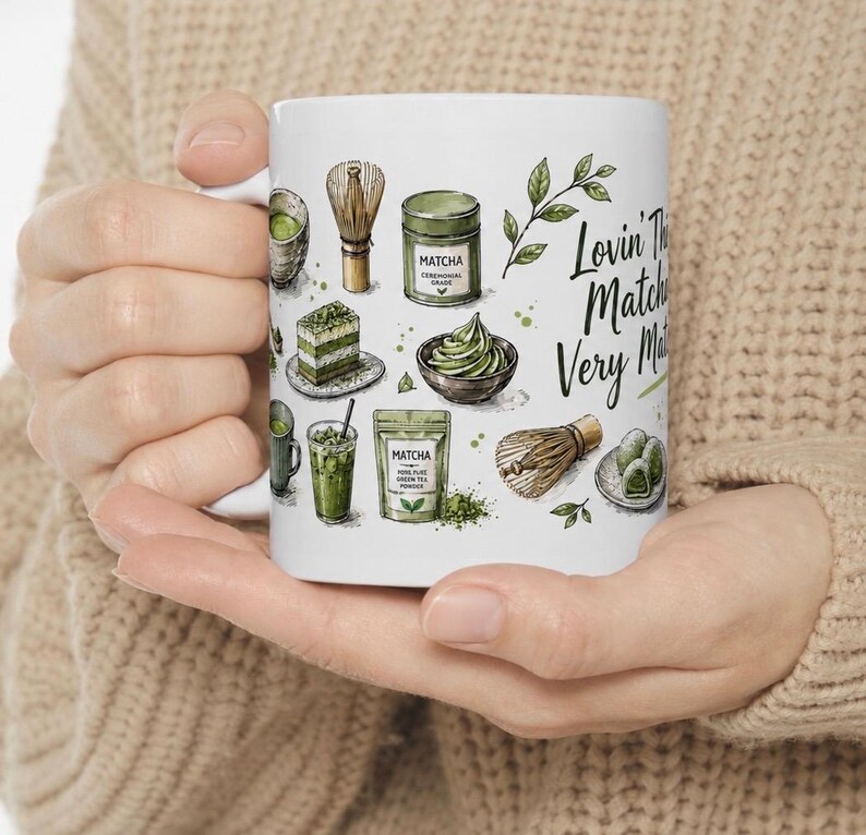May include: White ceramic mug featuring matcha-themed illustrations, including a whisk, powder, and cake. The mug has the text "Lovin' The Matcha Very Matcha" in a handwritten style. The mug is held by a person wearing a beige sweater.