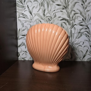 Może przedstawiać: Brzoskwiniowy ceramiczny wazon w kształcie muszli. Wazon ma żebrowaną fakturę i błyszczące wykończenie. Podstawa ma solidny, zaokrąglony kształt. Tło przedstawia wzór tapety w kwiaty.