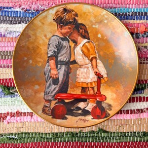 May include: A round decorative ceramic plate with a gold-toned rim featuring a painted illustration of two children leaning toward each other. One child wears a grey outfit, the other a white and yellow dress, both standing by a bright red toy scooter.