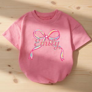 May include: A pink short-sleeved sweatshirt featuring a colorful bow design with the name "Emily" in a script font. The bow has rainbow-colored accents, and the sweatshirt is made of a soft material.