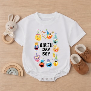 May include: White baby onesie with a colorful graphic of cartoon characters and the text "BIRTH DAY BOY". The onesie is surrounded by a wooden bunny rattle, a wooden rainbow, and brown baby shoes.