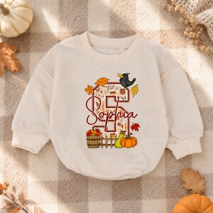 May include: A cream-colored long-sleeve sweatshirt featuring a large letter S filled with autumn leaf patterns. The design includes a black crow, a wooden barrel of apples, a green pear, and an orange pumpkin. The name Sophia is written in red script.