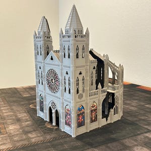 May include: A detailed, gray miniature cathedral model with two spired towers and a rose window. The model features intricate architectural details, including arched windows and buttresses. The cathedral has a section that appears damaged, revealing interior details. It sits on a patterned surface.