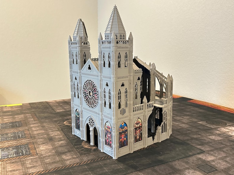 May include: A detailed, gray miniature cathedral model with two spired towers and a rose window. The model features intricate architectural details, including arched windows and buttresses. The cathedral has a section that appears damaged, revealing interior details. It sits on a patterned surface.