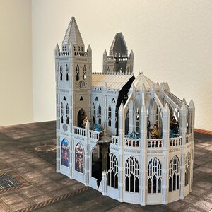 May include: A detailed, white miniature cathedral with multiple spires, arched windows, and decorative elements. The model features a complex design with a partially open structure, showcasing interior details and figures. The cathedral sits on a patterned surface.