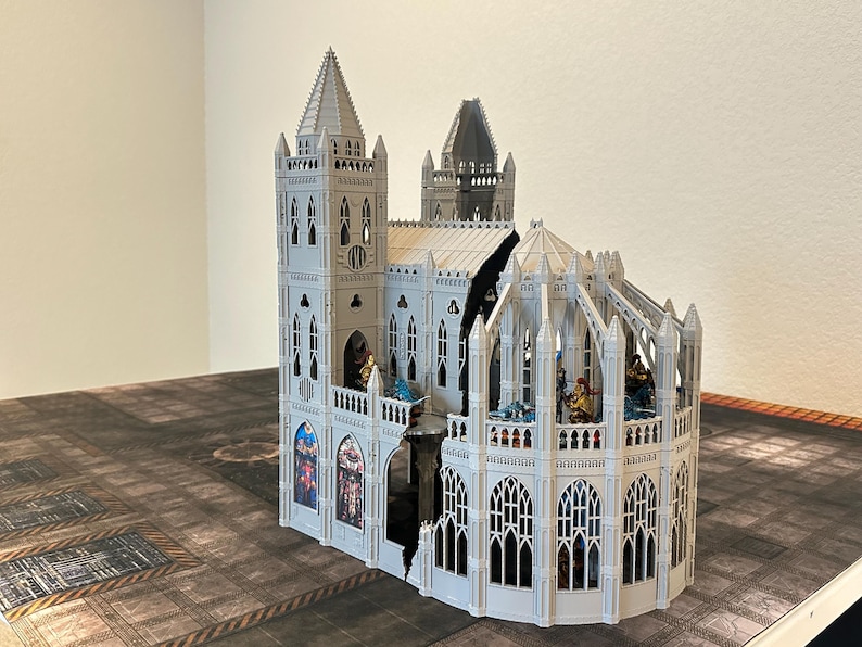 May include: A detailed, white miniature cathedral with multiple spires, arched windows, and decorative elements. The model features a complex design with a partially open structure, showcasing interior details and figures. The cathedral sits on a patterned surface.