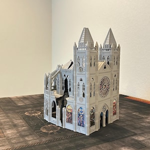 May include: A detailed, light gray model of a cathedral with two spired towers, arched windows, and a large rose window. The model features intricate architectural details and colorful stained-glass window designs. It is set on a textured, dark surface.