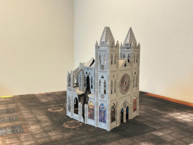 May include: A detailed, light gray model of a cathedral with two spired towers, arched windows, and a large rose window. The model features intricate architectural details and colorful stained-glass window designs. It is set on a textured, dark surface.