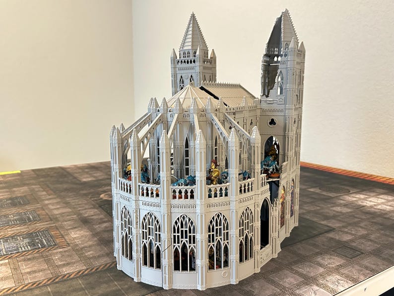 May include: A detailed, white miniature cathedral model with multiple spires, arched windows, and intricate architectural details. The model is partially assembled and sits on a patterned surface, suggesting a tabletop game setting.
