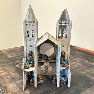 May include: A gray miniature cathedral model with two tall towers and a central arched section. The model has detailed architectural features and is populated with small figurines. The cathedral sits on a patterned surface.