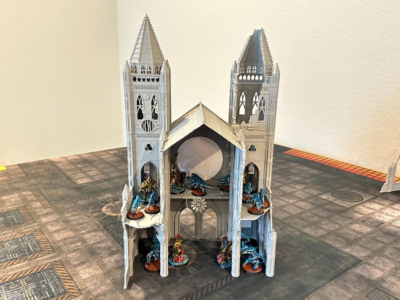 May include: A gray miniature cathedral model with two tall towers and a central arched section. The model has detailed architectural features and is populated with small figurines. The cathedral sits on a patterned surface.