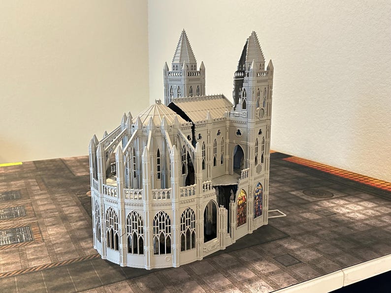 May include: A detailed, gray miniature cathedral model with intricate architectural details, including spires, arched windows, and decorative elements. The model is set on a textured surface, suggesting a tabletop game setting.