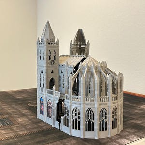 May include: A detailed, light gray model of a cathedral with multiple spires, arched windows, and intricate architectural details. The model is set on a textured surface, showcasing its gothic design.