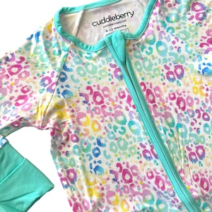 Puede incluir: Un pelele de bebé de manga larga turquesa y blanco con cierre de cremallera. El pelele tiene un estampado de leopardo acuarela colorido. La etiqueta dice "cuddleberry" y "6-12 meses".