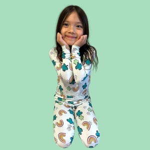 May include: A child's white pajama set featuring a rainbow, shamrock, and flower design. The long-sleeved top and trousers are patterned with colorful illustrations. The child is kneeling and smiling, with hands under their chin.