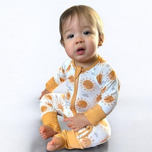 May include: A baby wearing a white and yellow long-sleeved onesie with a sun and cloud pattern. The onesie has a zipper down the front and a zipper at the ankles.