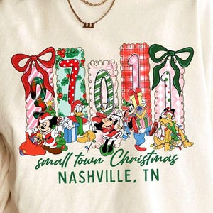 May include: Cream-colored t-shirt featuring a festive design with the text "2021" in colorful gift box shapes, each adorned with bows. Cartoon characters are gathered below. The text "small town Christmas Nashville, TN" is printed in green.