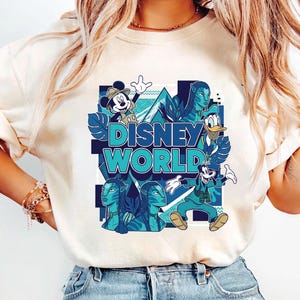 Puede incluir: Camiseta color crema con un vibrante gr&aacute;fico de Disney World. El dise&ntilde;o incluye a Mickey Mouse, el Pato Donald y personajes de Avatar, todos en tonos azules y turquesas. Las palabras "Disney World" se muestran de forma destacada.