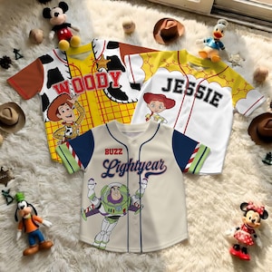 May include: Three baseball-style jerseys featuring characters from Toy Story. The jerseys are cream, yellow, and white with character graphics and names: Woody, Jessie, and Buzz Lightyear. The Buzz Lightyear jersey is cream with blue sleeves.