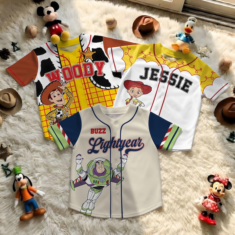 May include: Three baseball-style jerseys featuring characters from Toy Story. The jerseys are cream, yellow, and white with character graphics and names: Woody, Jessie, and Buzz Lightyear. The Buzz Lightyear jersey is cream with blue sleeves.