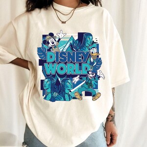 Puede incluir: Camiseta color crema con un dise&ntilde;o gr&aacute;fico. El dise&ntilde;o presenta el texto "DISNEY WORLD" en turquesa y azul, junto con ilustraciones de Mickey Mouse, el Pato Donald, Goofy y personajes de Avatar.